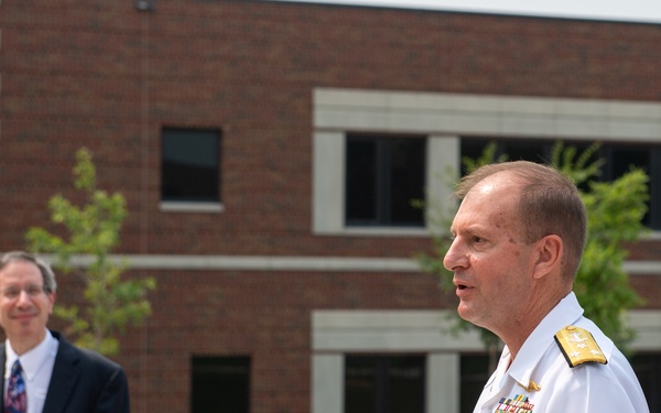 A senior U.S. Navy officer in white dress uniform with multiple service ribbons and gold shoulder boards is speaking at a microphone during an outdoor ceremony. A civilian man in a dark suit and patterned tie stands in the background, holding papers and a face mask. The setting appears to be outside a multi-story brick institutional or government building.
