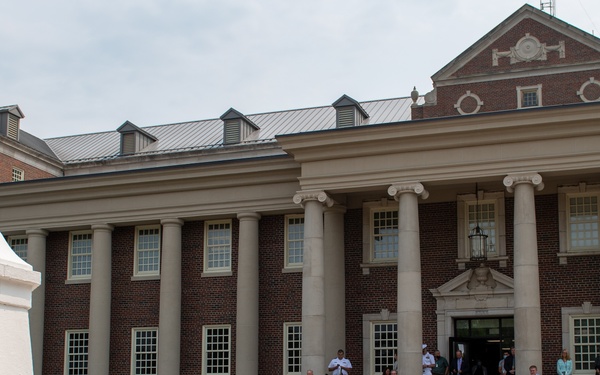 A military ensemble in white dress uniforms performs outdoors in a brick courtyard in front of a large Georgian-style red brick building with white columns. A crowd of spectators watches from chairs and the building's steps, suggesting a formal ceremony or public event. The setting appears to be a government or military installation based on the architecture and uniforms.