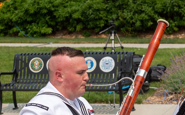 A U.S. Navy sailor in dress white uniform is seated outdoors on a brick plaza playing a bassoon. The uniform patch reads 'Navy Band Great Lakes,' indicating this is a performance by the Navy Band Great Lakes ensemble. The setting appears to be an outdoor military or public venue with military branch emblems visible on a nearby bench.