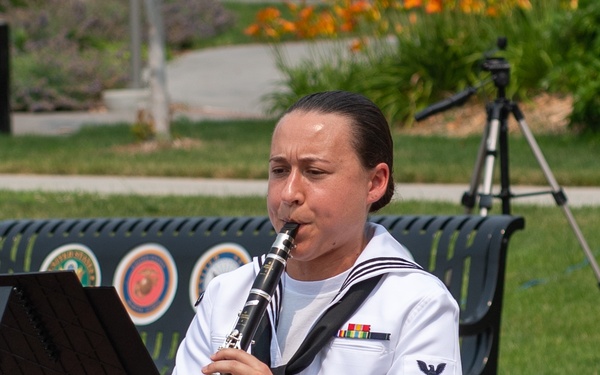 A female U.S. Navy enlisted musician in Service Dress White uniform is seated on a black chair playing a clarinet outdoors on a brick plaza. She is reading sheet music from a black music stand and wears ribbons and a Petty Officer insignia on her left sleeve. A bench with military branch emblems, including the Marine Corps seal, is visible in the background.