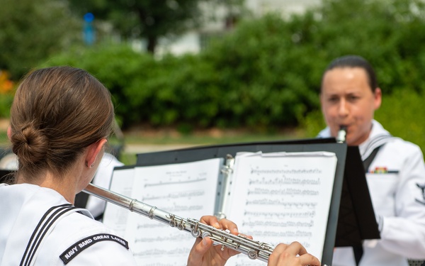 Two U.S. Navy musicians in white dress uniforms perform outdoors, reading from sheet music on stands. The foreground musician plays a silver flute, while the background musician plays a clarinet. A patch on the foreground sailor's uniform reads 'Navy Band Great Lakes.'