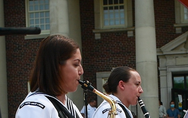 Two U.S. Navy musicians in white dress uniforms are seated outdoors performing on a brick plaza in front of a red brick building with white columns. The foreground musician plays an alto saxophone while the background musician plays a clarinet. Onlookers, some wearing medical scrubs and face masks, are visible in the background.