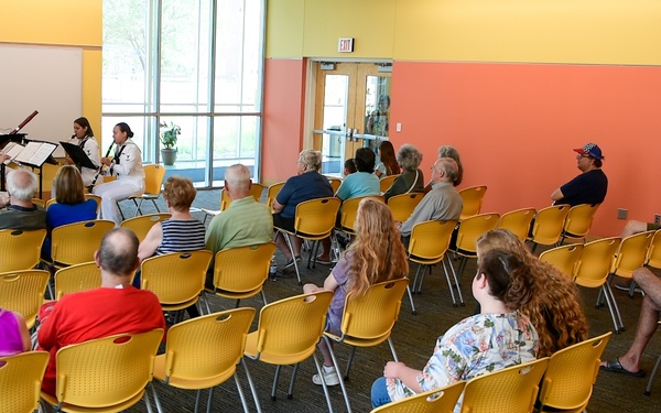 A small chamber music ensemble in white military dress uniforms performs for a sparse audience in a colorful community or public building. The performers appear to be woodwind players, likely from a military band, reading from music stands. The audience of approximately 20-25 people is seated in yellow plastic chairs arranged in rows, with many seats remaining empty.
