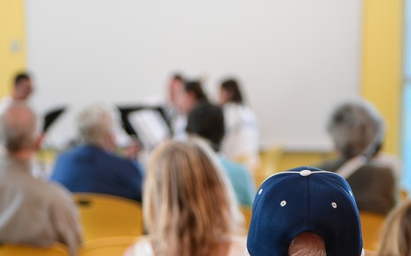A group of people are seated in yellow chairs attending what appears to be a presentation or small performance in a bright, yellow-walled room. The foreground shows a man wearing a blue 'NAVY' baseball cap and blue t-shirt, viewed from behind. At the front of the room, several individuals in white coats and dark clothing appear to be presenting or performing with music stands visible.