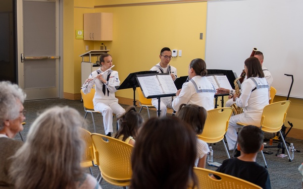 A small chamber music ensemble of approximately five musicians in white naval uniforms performs for a mixed audience of adults and children in a community room or library setting. The performers are seated in a semicircle with music stands, playing woodwind and brass instruments. The audience is seated on yellow plastic chairs arranged in rows facing the performers.