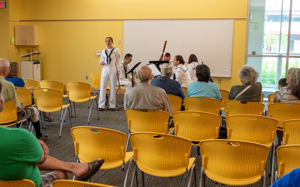 A small musical performance or presentation is taking place in a bright, modern community or institutional room with yellow walls and chairs. Navy personnel in white uniforms are standing at the front, with one appearing to speak or perform near a music stand. A modest audience of mostly older adults is seated in yellow chairs, with many seats remaining empty.