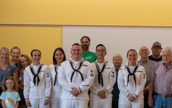 A group photo taken in what appears to be a community center or public building, featuring four U.S. Navy sailors in white dress uniforms surrounded by a large group of civilians of various ages. The gathering appears to be a military recognition or promotion ceremony, with family members and supporters present. The setting includes yellow chairs arranged in rows in the foreground and a whiteboard visible in the background.