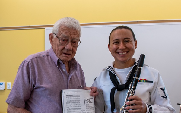 An elderly man and a female U.S. Navy musician pose together in what appears to be a community or meeting room. The sailor is holding a clarinet and wearing a white dress uniform with ribbons and a Petty Officer insignia. The elderly man is holding an open magazine or booklet featuring an article titled 'A First Time for Everything' with a black-and-white group photo visible.