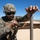 Talisman Sabre 21: U.S., Australian forces perform bilateral fuel training A U.S. Marine in full combat gear is working to set up or assemble a tan/desert-colored military tent or shelter structure in an open, arid outdoor environment. The soldier is wearing MARPAT digital camouflage, a tactical helmet with NVG mount, ballistic goggles, and a plate carrier vest. The setting appears to be a training area or forward operating base in a desert or semi-arid region under clear blue skies.