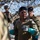 Talisman Sabre 21: U.S., Australian forces perform bilateral fuel training Military personnel are engaged in what appears to be a training exercise under camouflage netting in an outdoor setting. The central figure, wearing combat uniform and protective goggles pushed up on his forehead, is being assisted or examined by another soldier wearing teal/turquoise chemical-resistant gloves. Additional uniformed personnel are visible in the background.