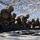 Talisman Sabre 21: U.S., Australian forces perform bilateral fuel training A group of military personnel in combat uniforms and helmets are working together to handle or fold a large dark material, likely a tarp or equipment cover, on a flat surface. The scene takes place beneath a camouflage netting canopy in what appears to be an arid, outdoor training or operational environment. A supervising figure in different attire stands in the background observing the activity.