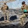 Talisman Sabre 21: U.S., Australian forces perform bilateral fuel training Military personnel in camouflage uniforms are engaged in what appears to be a field training exercise or base setup activity. One soldier in a distinctive green camouflage pattern is actively pulling a rope or strap while wearing protective goggles and teal-colored gloves. A second soldier in desert MARPAT camouflage with body armor and helmet stands observing in the background.