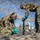 Talisman Sabre 21: U.S., Australian forces perform bilateral fuel training Two military personnel in camouflage uniforms and teal/green protective gloves are working with what appears to be a mortar tube on a sandy surface beneath a camouflage net. A group of additional uniformed soldiers observes in the background. The setting appears to be a training exercise conducted outdoors in a desert or sandy environment.