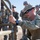 Talisman Sabre 21: U.S., Australian forces perform bilateral fuel training A group of military personnel in camouflage uniforms are gathered outdoors in what appears to be an arid environment. The foreground soldier, wearing a boonie hat and sunglasses, is actively demonstrating or working on a piece of military equipment with metal tubing/frame. The background soldiers, wearing different camouflage patterns suggesting a joint or multi-branch exercise, are observing attentively.