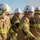 Talisman Sabre 21: U.S., Australian forces perform bilateral fuel training Four firefighters in full protective gear are engaged in what appears to be a firefighting operation or training exercise in an open, arid outdoor environment. They are wearing tan/gold turnout gear with high-visibility yellow reflective stripes and white helmets with face shields. A water spray is visible on the right side of the image, suggesting active hose deployment.