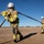 Talisman Sabre 21: U.S., Australian forces perform bilateral fuel training Two firefighters in full protective gear are pulling a large hose across an open, dry dirt field under a clear blue sky. The foreground firefighter is leaning forward with significant physical effort, gripping the hose with both gloved hands. The setting appears to be a training exercise or drill in a rural or semi-arid environment.