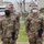 Four U.S. Army soldiers in OCP (Operational Camouflage Pattern) uniforms are standing outdoors on what appears to be a military installation, likely in South Korea based on visible Korean signage and architectural style. Two soldiers in the foreground are wearing face masks and holding dark-colored folders or booklets, suggesting a formal ceremony or official event. Name tapes visible include 'GRIMMAGE' and 'MERCADO' on the front soldiers, with 'GARCIA' partially visible.
