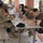 A group of U.S. Army soldiers and civilians are eating together at a picnic table under an outdoor covered pavilion, suggesting a unit social event or cookout. Several individuals are wearing U.S. Army OCP (Operational Camouflage Pattern) uniforms with American flag patches, while others are in civilian attire. The setting appears to be on a military installation based on the brick structures and overall environment.