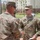 Two U.S. Army soldiers in OCP (Operational Camouflage Pattern) uniforms are engaged in what appears to be an award or recognition ceremony outdoors on a military installation. The soldier facing the camera is identified by his name tape as 'THOMSEN' and wears U.S. Army insignia. A third soldier is partially visible in the background.