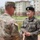 A U.S. Army soldier and a Republic of Korea Army (ROKA) soldier are engaged in a handshake or exchange on what appears to be a military installation. The U.S. soldier is wearing OCP (Operational Camouflage Pattern) uniform with an American flag patch and unit insignia, while the ROKA soldier wears a distinctive black beret and digital camouflage uniform with 'ROKA' and Korean text visible. The setting appears to be Camp Humphreys or a similar joint U.S.-South Korean military base.