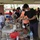 An outdoor buffet-style food event is taking place under a covered pavilion, likely at a military base given the presence of uniformed personnel and institutional buildings in the background. People are serving themselves from a long table filled with various food trays, chafing dishes, and snack items. Several attendees are wearing face masks, suggesting this was taken during or after the COVID-19 pandemic period.