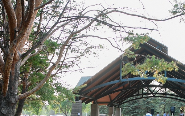 An outdoor community concert or performance event taking place at a park bandshell/pavilion during evening hours. Audience members are seated on folding chairs and lawn chairs across the grass, with some gathered under large pine trees in the foreground. Performers are visible on or near the covered stage structure.