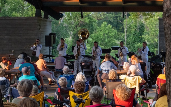 An outdoor concert is taking place at a park pavilion, where a military band — likely a U.S. Navy band based on the white uniforms — performs brass and wind instruments for a casual audience. Spectators of various ages are seated in lawn chairs and folding chairs on grass, enjoying the performance in a relaxed, community setting. The scene is framed by two large tree trunks in the foreground, with lush green trees visible in the background.