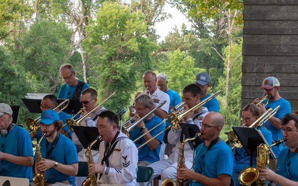 A large jazz or concert band called 'KICKS - Band of Fargo Moorhead' is performing outdoors at what appears to be a park pavilion or bandshell. The ensemble includes approximately 20 musicians, most wearing matching blue t-shirts, with a few members in white Navy uniforms. The performance features brass and woodwind instruments in a big band format.