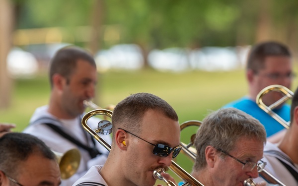 A group of musicians are playing trombones and brass instruments outdoors, likely at a public park performance. Several musicians are wearing white naval/sailor uniforms, while others wear teal/turquoise shirts. The image captures a mid-performance moment with multiple players actively playing their instruments.