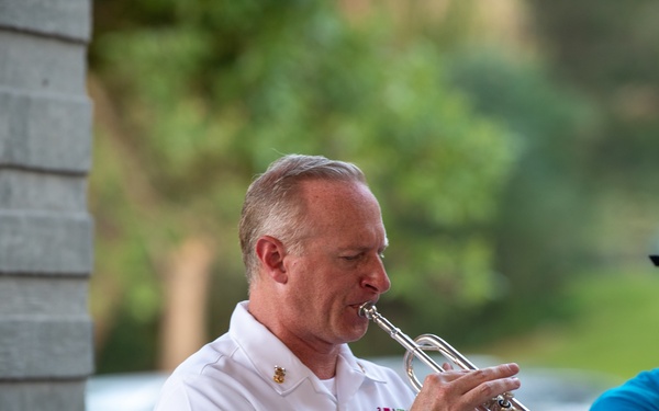 Two musicians are playing trumpets outdoors, likely at a public performance or community event. The central figure is wearing a white military dress uniform with ribbons and a name tag, suggesting an official or ceremonial context. A second musician in a blue t-shirt and gray cap is partially visible in the background.