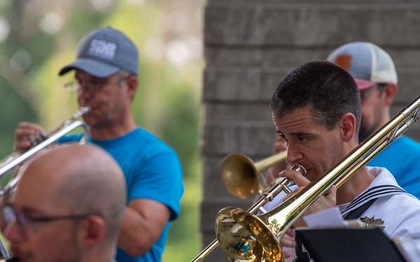 An outdoor musical performance featuring a brass and woodwind ensemble. Multiple musicians are playing instruments including trombone, trumpet, and saxophone, with one performer visibly wearing a white naval/military uniform with a black tie. The setting appears to be a park or outdoor pavilion with green trees visible in the background.