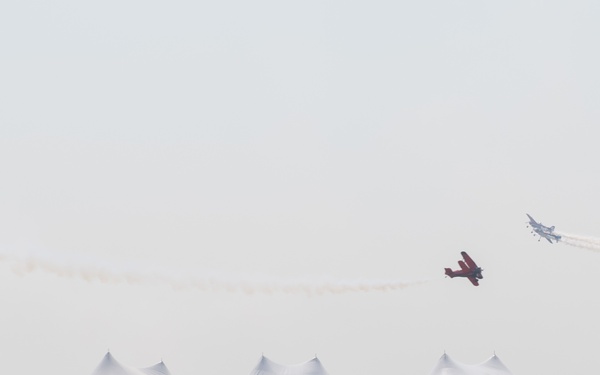A large outdoor airshow event with a substantial crowd of spectators watching aircraft perform aerial maneuvers. Multiple planes are visible in the sky, including what appears to be a biplane and at least one larger aircraft leaving smoke trails. The venue features numerous white event tents, orange crowd-control barriers, and colorful sponsor banners.