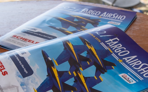 Two Fargo AirSho program booklets are displayed on a wooden table, both featuring the United States Navy Blue Angels on their covers. The programs are sponsored by RAD Aerosports and show advertisements from SCHEELS on the back cover. The booklets appear to be from the same event, likely held on the 24th and 25th of a given month.
