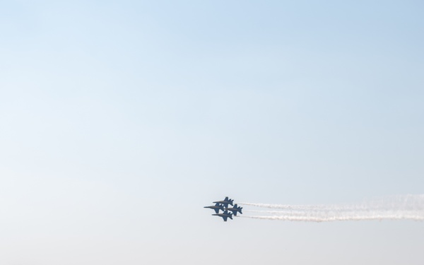 A crowd of spectators watches a military flight demonstration, likely performed by the U.S. Navy Blue Angels, at an airshow. Multiple jet aircraft are visible in tight formation with white smoke trails against a hazy sky. Audience members are photographing and filming the performance from behind an orange safety barrier fence.
