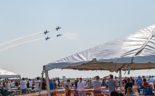 An outdoor airshow event with a large crowd gathered on a grassy field beneath white event tents. Four military jets, likely the Blue Angels or Thunderbirds, are performing a formation flyover leaving white contrails across the sky. Spectators of various ages are watching the aerial display, some standing and some seated in folding chairs.