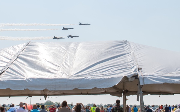 An outdoor airshow event with a large white event tent in the foreground and a crowd of spectators watching military jets fly in formation overhead. The jets are leaving white contrails across a clear blue sky. The setting appears to be at an airfield or open flat terrain with tables and chairs set up under the tent.