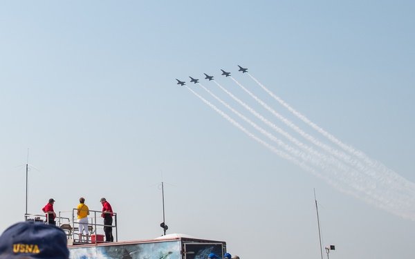 An air show event featuring a formation of jet aircraft leaving white contrail smoke trails across a clear sky. In the foreground, a man wearing a USNA (United States Naval Academy) cap photographs the aerial display. The ground area includes spectators, parked cars, a decorated trailer/stage with people on it, and orange safety fencing.