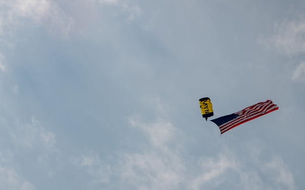 A skydiver with a small deployed parachute is descending through a partly cloudy sky while carrying a large American flag. The parachute appears to be yellow and black with text visible on it, consistent with a military demonstration team. This appears to be a patriotic aerial demonstration, likely at an airshow or public event.