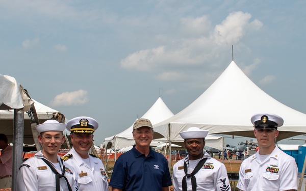 Five individuals pose for a group photo at an outdoor event, likely an airshow or military public event. Four of the individuals are wearing U.S. Navy uniforms in white dress attire, including one officer with a gold-braided cover, while the fifth person in the center is a civilian wearing a navy blue polo shirt and khaki pants. White event tents and what appears to be a military aircraft are visible in the background.