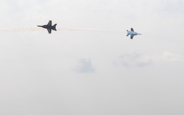 An air show is taking place at what appears to be a military airfield, with two jet aircraft — one in Blue Angels livery and one in a gray camouflage scheme — performing a crossing maneuver while trailing smoke. Spectators, including uniformed Navy personnel and civilians, watch from a designated viewing area with tables and chairs. The scene suggests a formal or VIP viewing section at a military aviation event.