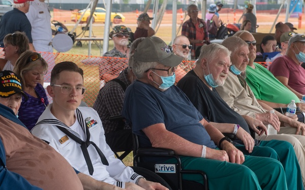A group of military veterans and a Navy sailor are seated together at an outdoor event, likely a veterans' ceremony or air show. Several attendees are in wheelchairs and wearing hospital wristbands, suggesting they may be patients from a VA medical facility. The gathering appears to be held under a tent with a crowd visible in the background.