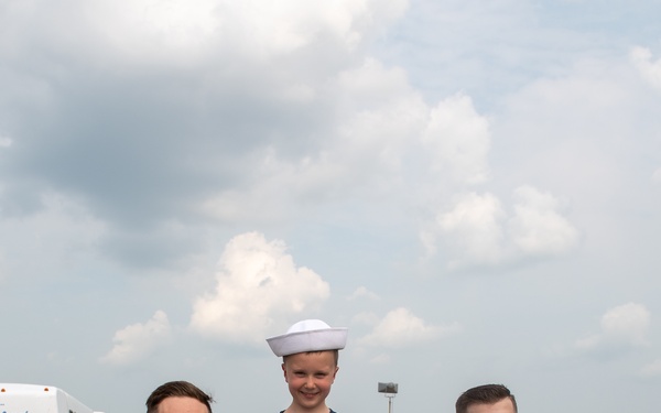Two U.S. Navy sailors in white dress uniforms are crouching down on either side of a young child at what appears to be an outdoor public event or air show. The child is wearing a navy blue American flag t-shirt and has been given one of the sailor's white dixie cup hats to wear. The scene conveys a friendly community outreach or public relations moment.