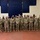 A group of approximately 18-20 military personnel in U.S. Army OCP (Operational Camouflage Pattern) uniforms are posed together for a group photo inside what appears to be a military facility or armory. All individuals are wearing face masks, consistent with COVID-19 pandemic protocols. The group includes both male and female soldiers standing in two rows.