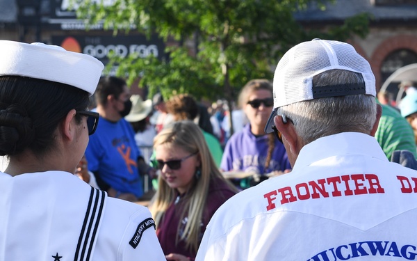 Two individuals are seen from behind at what appears to be the Cheyenne Frontier Days event. One person is wearing a U.S. Navy sailor uniform with a white dixie cup hat, while the other wears a white Western-style shirt advertising 'Frontier Days Chuckwagon Breakfasts' by Kiwanis International. They are surrounded by a crowd of attendees at an outdoor public event.