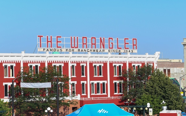 A large outdoor festival or public event is taking place in what appears to be a downtown area, with hundreds of attendees gathered in an open plaza. The prominent backdrop is 'The Wrangler' building, a red brick structure with a rooftop sign reading 'Famous for Ranchwear Since 1943.' The crowd is densely packed, with many attendees wearing cowboy hats, suggesting a Western-themed or rodeo-related event.