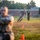 Officer Candidates School Combat Fitness Test Military personnel, appearing to be U.S. Marines based on visible camouflage uniforms and a green 'USMC' shirt, are conducting a physical fitness or combat readiness drill on a grassy field. Orange cones mark a course or lane, and a large group of uniformed personnel observes in the background. The foreground subject is blurred due to shallow depth of field, while the mid-ground runner is in sharp focus.