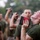 Officer Candidates School Combat Fitness Test A group of individuals in olive green military-style t-shirts are participating in what appears to be a military training exercise or drill. The foreground subject has the number '242' written on his hand and is tilting his head back while holding a red and black object up to his mouth. Multiple similarly dressed individuals are visible in the background performing the same action.