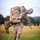 Officer Candidates School Combat Fitness Test Two individuals in U.S. Marine Corps desert MARPAT camouflage uniforms are performing a fireman's carry drill on a grass field. One service member is carrying the other on his back while running along a marked lane indicated by a white rope or line on the ground. The carrier displays visible physical exertion with a strained expression, suggesting a timed physical fitness or combat casualty evacuation exercise.