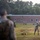 Officer Candidates School Combat Fitness Test Military personnel, identifiable as U.S. Marines based on visible 'USMC' text on a green shirt, are conducting a physical training exercise on a grassy field. One Marine in camouflage uniform is throwing or launching an object (likely a medicine ball or similar training equipment) while an instructor in a green USMC shirt observes closely. The scene takes place near a brick building on what appears to be a military installation.