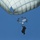 A parachutist is descending under two visible parachutes against a clear blue sky. The image is taken from a low angle looking upward, showing a large main canopy and a smaller reserve or drogue chute between the jumper and the main canopy. The jumper appears to be in full gear, silhouetted against the bright sky.